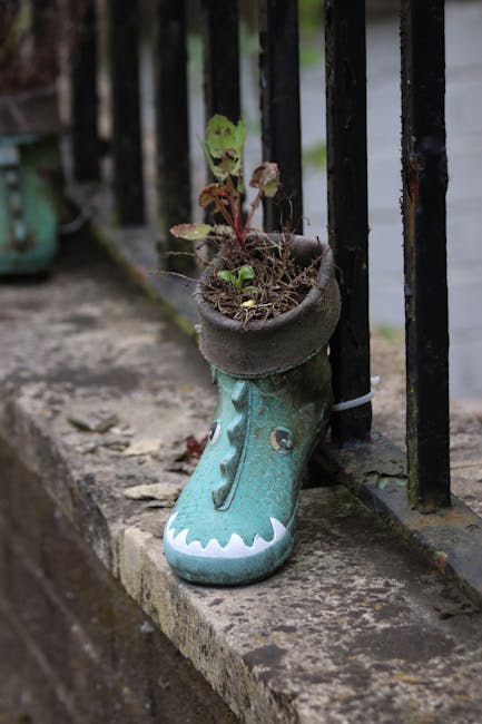 A close-up of an upturned decorative green rubber boot with a crocodile face design, repurposed as a plant pot containing small leafy plants and soil. The boot sits on a concrete pavement next to a bl